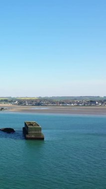 Asnelles, Normandiya 'daki Gold Sahili yakınlarında açık mavi gökyüzünün altında, uzak bir köy ve kumlu bir kıyı şeridi bulunan yapay liman yapısının havadan görüntüsü..