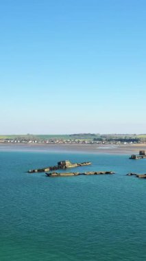 Asnelles, Normandiya kıyısında sakin mavi sularda, kıyı köyü ve arka planda açık gökyüzü olan yapay bir liman yapısının kalıntıları..