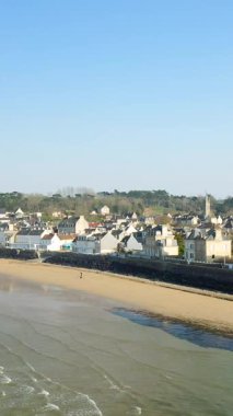 Arromanches-les-Bains 'i, kumlu bir sahil boyunca geleneksel evleri ve açık mavi gökyüzünün altında hafif dalgaları geniş açıdan görebiliriz..