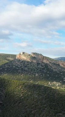 Engebeli bir kayalık tepe, parçalı bulutlu bir gökyüzünün altındaki çalı kaplı yamaçların üzerinde yükselir. Doğal arazi dramatik ve huzurlu bir kırsal alan yaratır..