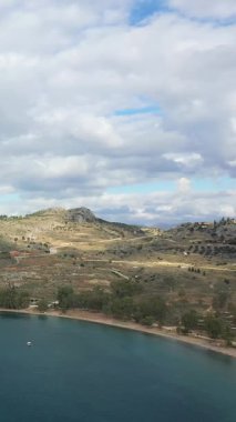 Yunanistan 'ın Mora kırsalında, parçalı bulutlu bir gökyüzünün altında yuvarlanan tepelerin manzarası ve sakin bir koy. Dolambaçlı yolları ve seyrek bitkileri olan doğal manzara..