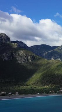 Yunanistan 'ın Monemvasia kenti yakınlarındaki yemyeşil dağlarla turkuaz kıyı şeridi manzarası ve dağınık evler parçalı bulutlu bir gökyüzü altında.