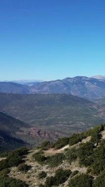 Açık mavi gökyüzünün altındaki engebeli yeşil dağların geniş bir görüntüsü, uzak bir tepenin üzerinde rüzgar türbinleri görülebiliyor. Sürdürülebilirlik ve sükunet kavramları için ideal doğal, manzaralı ortam.