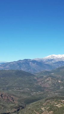 Açık, güneşli bir günde, yuvarlanan yeşil tepelerin üzerinde yükselen karlı dağların geniş manzarası. Manzarada dağınık kırsal evler ve dolambaçlı yollar yer alıyor..