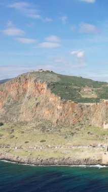 A dramatic rocky cliff rises above the historic medieval town of Monemvasia on the coast of Greece, with turquoise sea in the foreground and blue sky above.