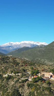 Yunanistan 'da açık mavi bir gökyüzü altında, yemyeşil tepeler ve uzak karlı dağlarla çevrili tarihi Osios Loukas Manastırı' nın havadan görünüşü.