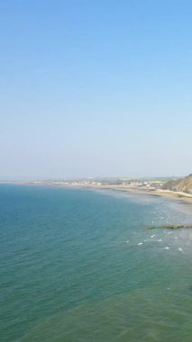 Arromanches-les-Bains 'in sahil şeridini ve kumlu plajını açık mavi gökyüzünün altında geniş bir görüş açısına sahip. Dalgalar kıyıya yaklaşırken uzak kayalıklar ve deniz kıyısındaki köy görünür..