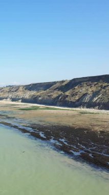 Normandiya 'nın Port en Bessin kentindeki açık mavi gökyüzünün altında engebeli uçurumların ve kumlu kıyı şeridinin manzarası. Kıyı manzarasında kaya oluşumları ve sakin gelgit suları yer almaktadır..