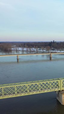 Kış sahnesinde Sully-sur-Loire 'daki Loire Nehri' nin üzerinde karla kaplı ağaçlar ve berrak bir gökyüzü altında sakin bir su bulunan metal bir köprü var..