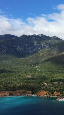 Yunanistan 'ın Monemvasia kenti yakınlarındaki turkuvaz suları, yemyeşil yemyeşil yemyeşil ve dramatik dağlarla dolu engebeli bir kıyı şeridi manzarası parçalı bulutlu bir gökyüzü altında.