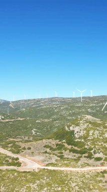 Yunanistan 'ın Thisbe kenti yakınlarındaki açık mavi gökyüzünün altındaki yeşil tepelerde bir sıra rüzgar türbini duruyor. Arazide kayalık arazi ve dolambaçlı toprak yollar yer alıyor ve Akdeniz ortamında yenilenebilir enerjiyi vurguluyor.