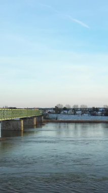 Fransa 'nın Sully sur Loire kentinde karla kaplı bankaları ve uzak evleri olan sakin bir nehri kapsayan yeşil bir köprüye sahip sakin bir kış manzarası..