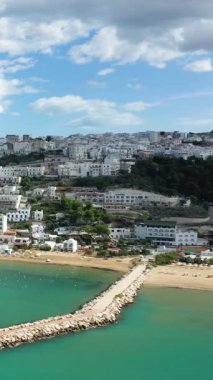 Peschici 'nin turkuaz sulara tepeden bakan beyaz tepe binaları ve parçalı bulutlu bir gökyüzünün altında kumlu bir plajı manzarası. Resimli İtalyan sahil kasabası yemyeşil ve taştan bir iskeleyle çevrili..