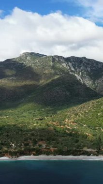 Yunanistan 'ın Monemvasia kenti yakınlarında turkuvaz suyu bulunan sakin bir plaja inen yemyeşil bir dağın manzarası. Dramatik bulutlar engebeli araziye gölge düşürür..