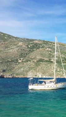 Beyaz bir yelkenli Porto Kagio 'nun berrak mavi sularında demirlemiş. Arkasında engebeli yeşil bir tepe var. Parlak bir gökyüzünün altında..