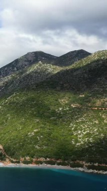 Yunanistan 'ın Monemvasia kenti yakınlarındaki bulutlu bir gökyüzünün altında kayalık bir kıyı şeridine ve turkuaz bir denize inen yeşil dağların manzarası.