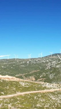 Yunanistan 'ın Thisbe kenti yakınlarındaki açık mavi gökyüzünün altındaki kayalık yeşil tepelerdeki rüzgar türbinlerinin geniş manzarası. Doğal arazi ve temiz teknolojiyle yenilenebilir enerji sahası.