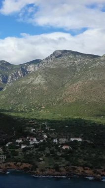 Yunanistan 'ın Monemvasia kenti yakınlarındaki küçük bir kıyı köyü üzerinde yükselen yeşil dağların manzarası, parçalı bulutlu bir gökyüzünün altında. Manzarada kayalık uçurumlar, yemyeşil bitki örtüsü ve mavi deniz yer alır..