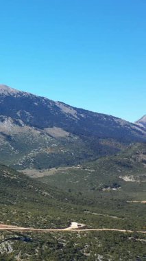 Yunanistan 'ın Thisbe kenti yakınlarındaki yeşil dağların ve açık mavi gökyüzünün geniş manzarası. Engebeli arazi ve dolambaçlı toprak yol bölgenin doğal güzelliğini ön plana çıkarıyor..