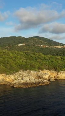 Yunanistan 'ın Syvota kentinde bulutlu bir gökyüzü altında yemyeşil tepeleri olan kayalık bir kıyı şeridi manzarası. Huzurlu kıyı manzarası güneş ışığıyla yıkandı..
