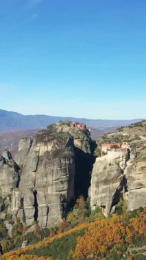 Renkli bir sonbahar manzarasının üzerinde yükselen etkileyici kayalık kayalıklar, açık mavi gökyüzünün altındaki zirvelerden birinin tepesinde duran tarihi bir manastır..