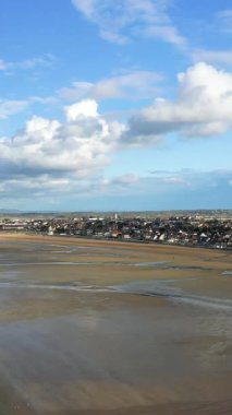 Lion-sur-Mer, Normandiya 'daki Sword Plajı' ndaki geniş kumlu sahil ve kıyı kasabası dağınık bulutlu mavi bir gökyüzünün altında. Gelgit desenleri ve uzak binalarla sakin deniz manzarası.