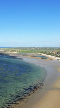 Kumlu kıyı şeridinin manzarası ve açık mavi sular. Fransa 'nın Grandcamp-Maisy şehrinde, parlak mavi bir gökyüzünün altında. Sahil boyunca görünen uzak tarlaları ve evleri olan sakin bir manzara..