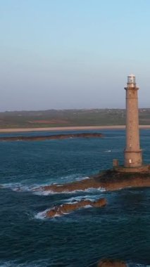 Uzun taştan bir deniz feneri, gün batımında açık bir gökyüzünün altında, Fransa 'nın Normandiya' nın Cap de la Hague kentinde denizle çevrili engebeli kayaların üzerinde duruyor..