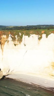 Etretat, Normandiya, Fransa 'da kumlu bir sahil ve açık bir gökyüzünün altında dramatik beyaz tebeşir uçurumları yükseliyor. Bereketli yeşil alanlar kayalıklar boyunca uzanıyor, çarpıcı bir kıyı manzarası yaratıyor..