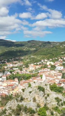Lauria, İtalya 'nın panoramik manzarasında, yeşil tepeler arasında yerleşmiş kırmızı çatıları olan geleneksel evler ve mavi gökyüzü altında bulutlu kayalık çıkıntılar görülüyor..