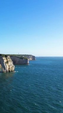 Etretat, Normandiya, Fransa 'da açık bir gökyüzünün altında mavi denizin üzerinde dramatik beyaz tebeşir kayalıkları yükseliyor. Doğal kaya oluşumları ve sakin okyanus sularıyla manzaralı kıyı manzarası.