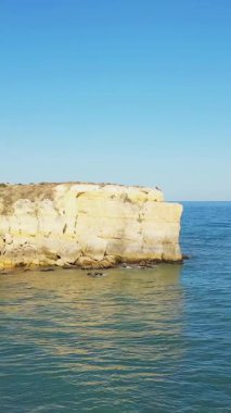 Portekiz 'in Albufeira kenti yakınlarındaki Algarve kıyısında berrak bir gökyüzünün altında, sakin mavi denizin üzerinde yükselen dramatik sarı kireçtaşı uçurumu. Engebeli doğal manzara seyahat ve doğa kavramları için ideal..