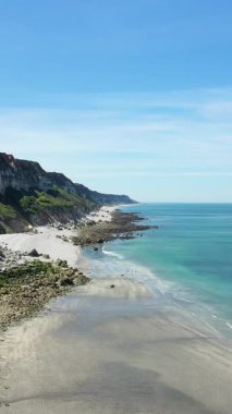 Berrak mavi gökyüzünün altında kumlu ve kayalık bir kıyı şeridi boyunca dramatik Normandiya uçurumları ve turkuaz denizin manzarası.