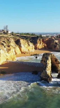Portekiz 'in Lagos yakınlarındaki Algarve sahilinde berrak mavi bir gökyüzünün altında engebeli deniz kayalıkları ve kumlu bir sahil manzarası..