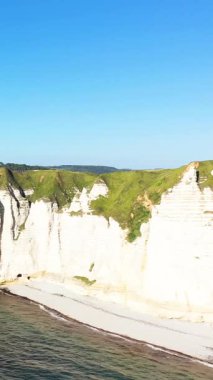Kumlu bir sahil ve açık bir gökyüzü altında mavi deniz üzerinde yeşil çimenli dramatik beyaz tebeşir uçurumları, Etretat, Normandiya, Fransa.