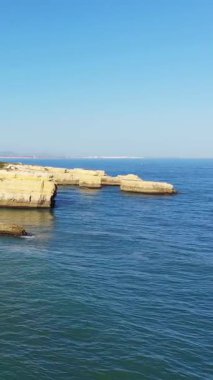 Portekiz, Albufeira yakınlarındaki Algarve kıyı şeridi boyunca uzanan engebeli sarı kayalıkların manzarası ve sakin mavi okyanus berrak bir gökyüzünün altında..
