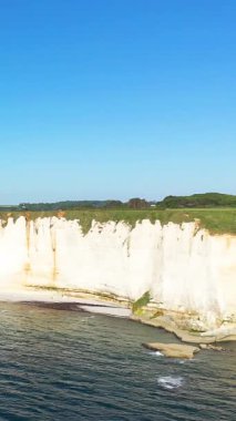 Etretat, Normandiya, Fransa 'da açık bir gökyüzü altında mavi denizin üzerinde dramatik beyaz tebeşir kayalıkları yükselir. Bereketli yeşillik kayalıkların tepesinde çarpıcı bir doğal manzara yaratıyor..