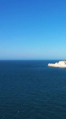 Fransa 'nın Normandiya kentinde açık bir gökyüzünün altında Etretat' ın ikonik beyaz tebeşir kayalıkları ile derin mavi okyanus manzarası. Doğal kaya oluşumları ve canlı renkleriyle sakin deniz manzarası.