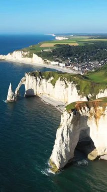 Fransa 'nın Normandiya kıyısındaki Etretat' taki ikonik beyaz tebeşir kayalıklarının ve doğal kemerlerin dramatik hava manzarası, açık bir gökyüzünün altında mavi deniz ve yeşil alanlar..