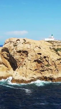 Korsika, Fanale di Petra 'daki derin mavi denize bakan engebeli uçurumların tepesinde beyaz bir deniz feneri duruyor. Parlak güneş ışığı dramatik kıyı manzarasını aydınlatır.