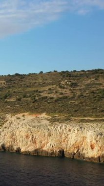 Dik kayalıklar, Yunanistan 'ın Mani bölgesindeki Diros plajı yakınlarında berrak bir gökyüzünün altında açık mavi denizin üzerinde yükseliyor. Engebeli araziyi kaplayan seyrek bitki örtüsü dramatik bir kıyı manzarası yaratıyor..