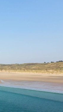 Plage de la Vieille Eglise 'de açık mavi gökyüzünün altında yumuşak dalgalar ve çimenli kumullarla geniş kumlu bir sahil. Huzurlu kıyı manzarası rahatlama ve doğa temaları için ideal.