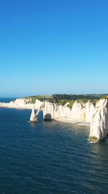 Fransa 'nın Normandiya kıyısındaki Etretat' ta açık bir gökyüzünün altında mavi denizin üzerinde dramatik beyaz tebeşir kayalıkları yükseliyor. İkonik doğal kemerler ve engebeli kıyı şeridi parlak güneş ışığında görülebilir..