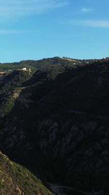 Korsika kırsalında açık mavi gökyüzünün altındaki engebeli yeşil dağların manzarası. Doğal güneş ışığı dramatik araziyi ve yemyeşil bitkileri vurguluyor.