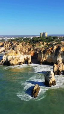 Portekiz 'in Albufeira kenti yakınlarındaki Algarve sahilindeki turkuaz okyanus dalgalarının üzerinde, açık mavi bir gökyüzünün altında dramatik kayalıklar yükseliyor..