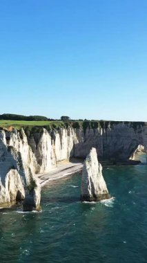 Etretat, Normandiya kıyıları boyunca açık mavi gökyüzünün altında turkuaz denizin üzerinde dramatik beyaz tebeşir kayalıkları yükselir. İkonik doğal kemerler ve kaya oluşumları çarpıcı bir kıyı manzarası yaratır..