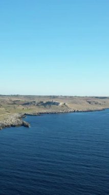 İtalya 'nın Porto Badisko şehrinde kayalık kıyı şeridi ve açık bir gökyüzünün altında derin mavi deniz manzarası. Manzarada engebeli uçurumlar ve Akdeniz kıyısı boyunca açık araziler yer alıyor..