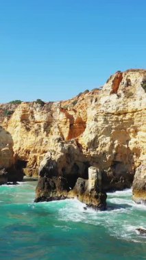 Portekiz 'in Lagos kenti yakınlarındaki Algarve kıyısındaki berrak turkuaz suların üzerinde parlak mavi bir gökyüzünün altında dramatik altın uçurumlar yükseliyor..