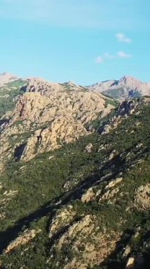 Korsika kırsalındaki açık mavi gökyüzünün altında engebeli kayalık dağların ve yemyeşil tepelerin manzarası. Dramatik arazi ve canlı bitki örtüsüyle doğal manzara.