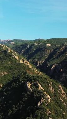 Korsika kırsalındaki engebeli yeşil dağlar ve kayalık uçurumlar berrak mavi bir gökyüzünün altında. Sahne, Korsika kırsalının doğal güzelliğini ve dramatik arazisini yakalıyor..